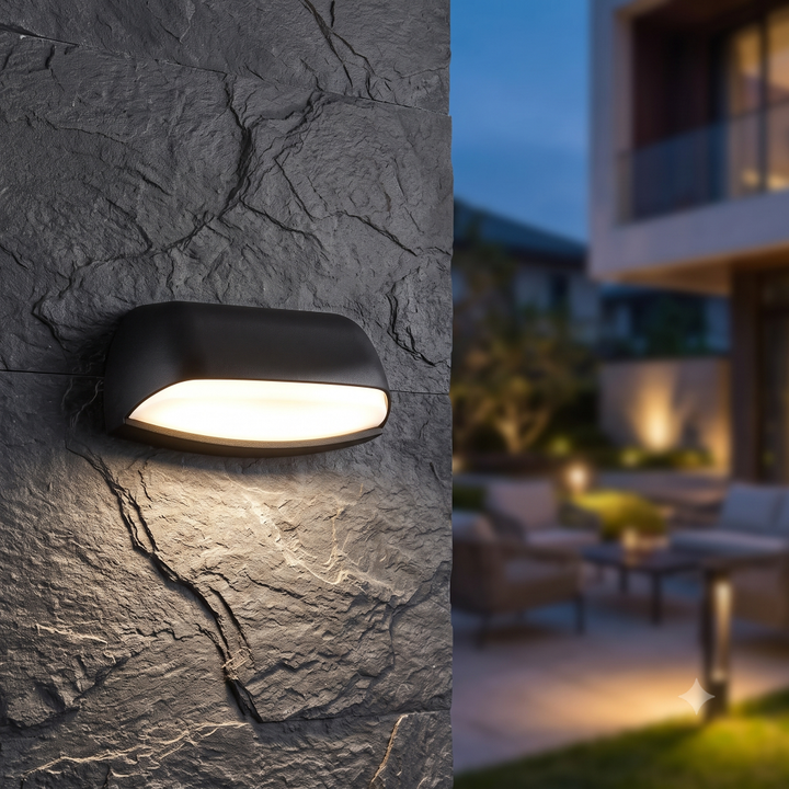 Modern zwarte entree buiten muurlamp Jessy met een diffuse lichtbundel naar beneden, gemonteerd op een stoere, donkere leistenen muur in een luxe sfeervolle patiotuin bij schemering.